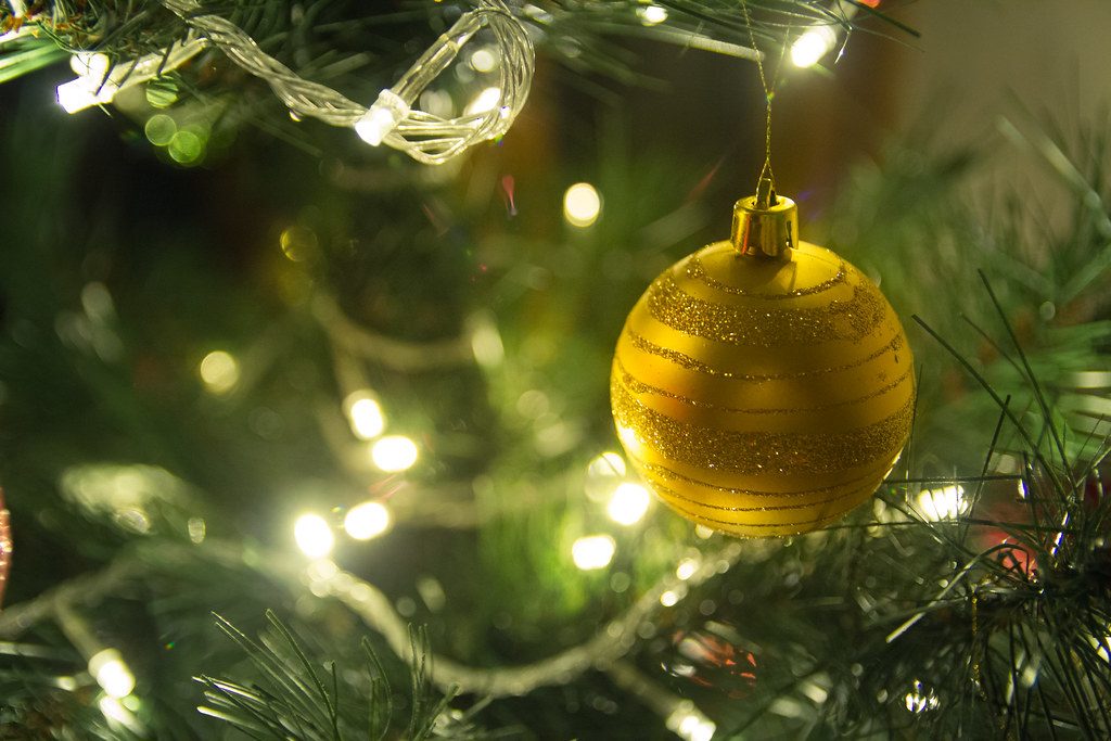 A gold glitter-striped bauble hanging on a Christmas tree among warm white fairy lights.