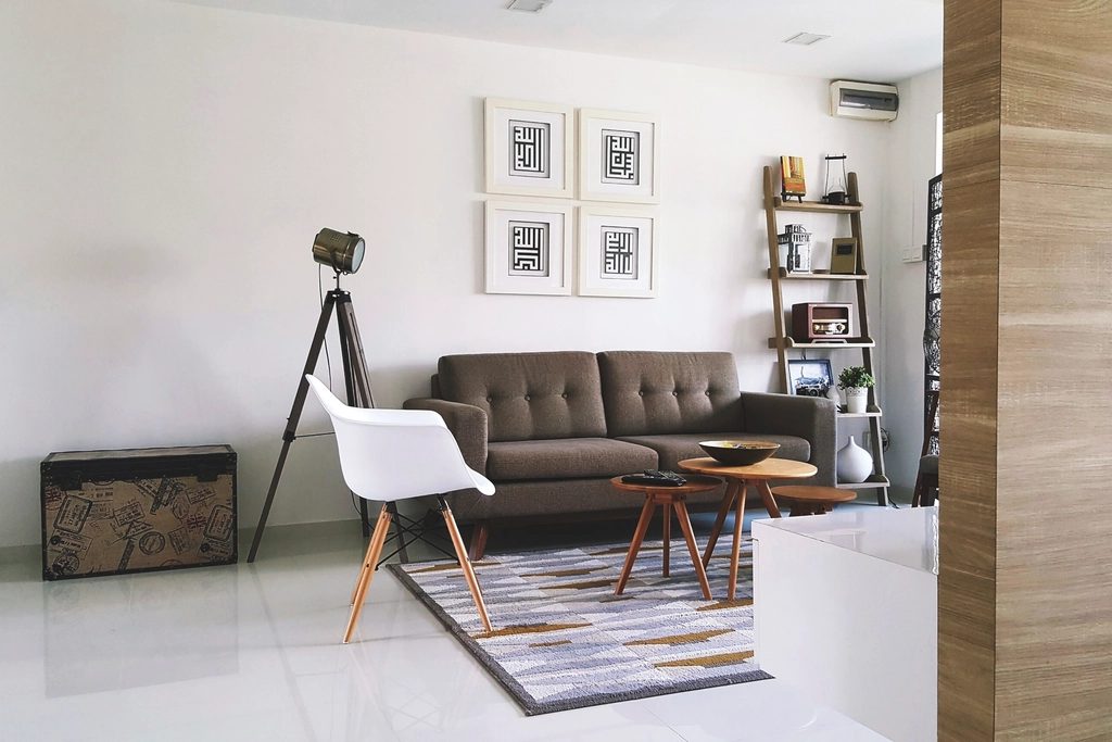 A bright living room: a grey-brown button-tufted sofa, a white moulded armchair on wooden legs, a spotlight floor lamp on a tripod, three round wooden side tables at three heights on a grey geometric rug, a leaning ladder shelf with a small retro radio, and a travel-print trunk.