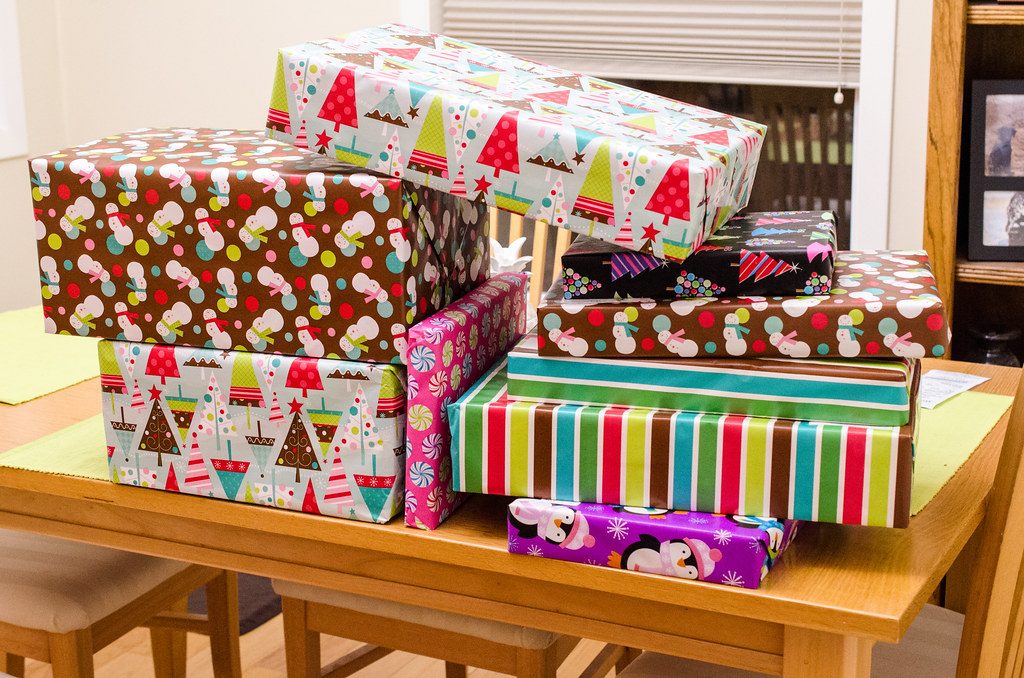 A stack of boxes in colourful holiday wrapping paper (snowmen, Christmas trees, stripes, penguins) on a wooden dining table.