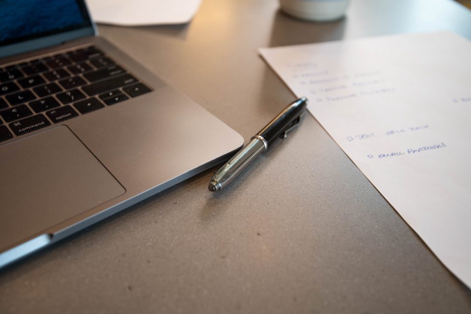 A silver laptop on a grey desk beside a pen and a sheet of handwritten notes.