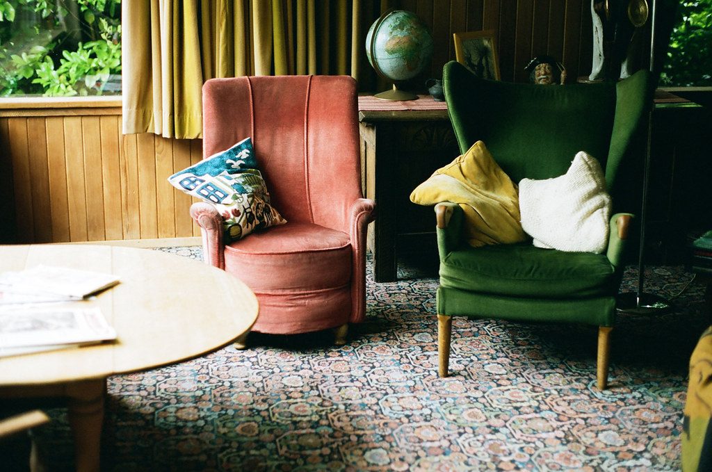 A styled living room with a coral velvet armchair and a green wingback chair with cushions, a globe on a side table, a patterned rug and a round wooden coffee table.