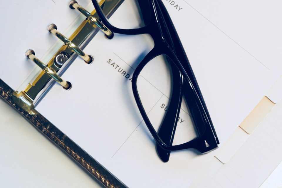 An open ring-bound planner at a week page with weekday headings and no dates, a pair of black glasses lying across it.