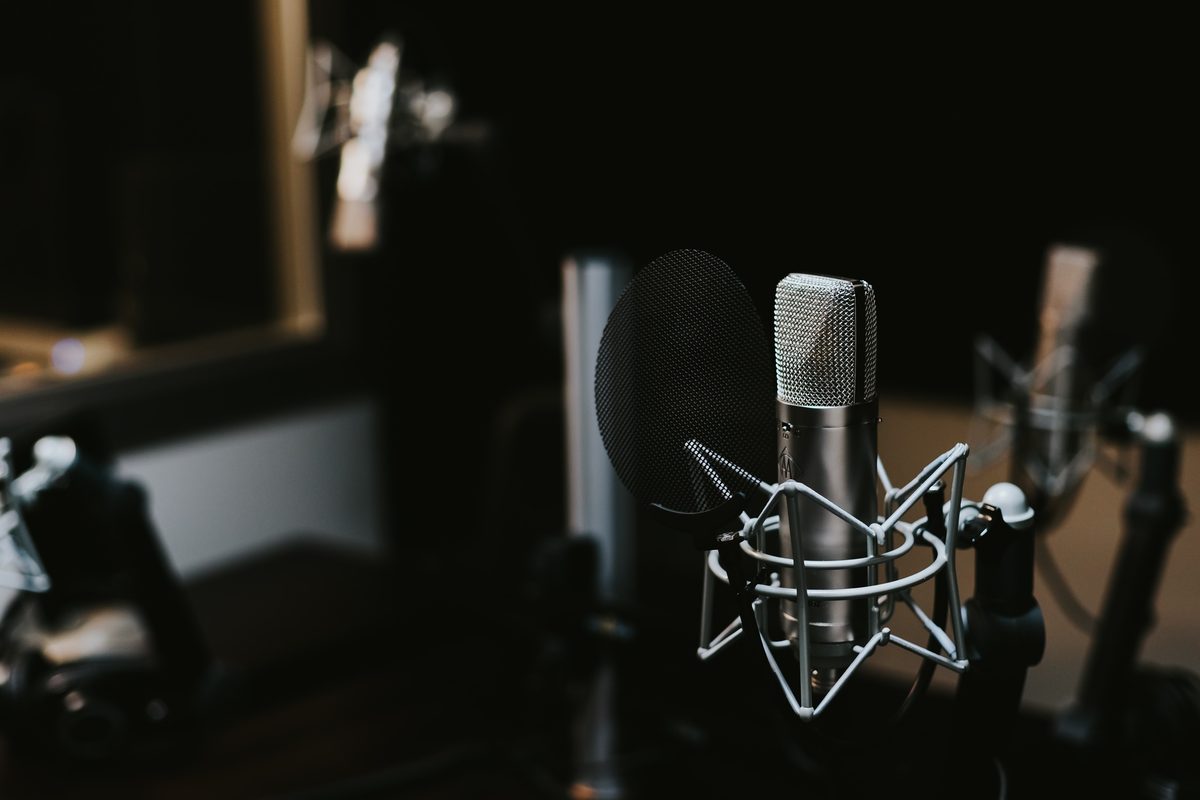 A silver studio condenser microphone in a shock mount with a pop filter, in a dim recording room.