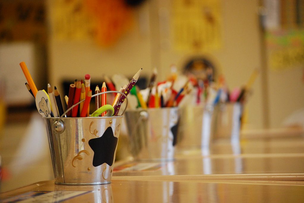 Small tin buckets filled with coloured pencils and pens lined up along a classroom table.