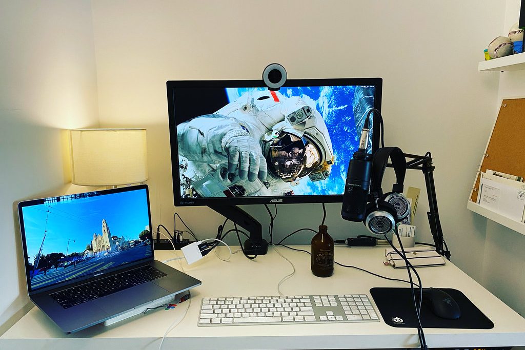 A home desk streaming setup with a monitor and ring webcam, a boom-arm microphone with headphones, a laptop, keyboard, mouse and a table lamp.