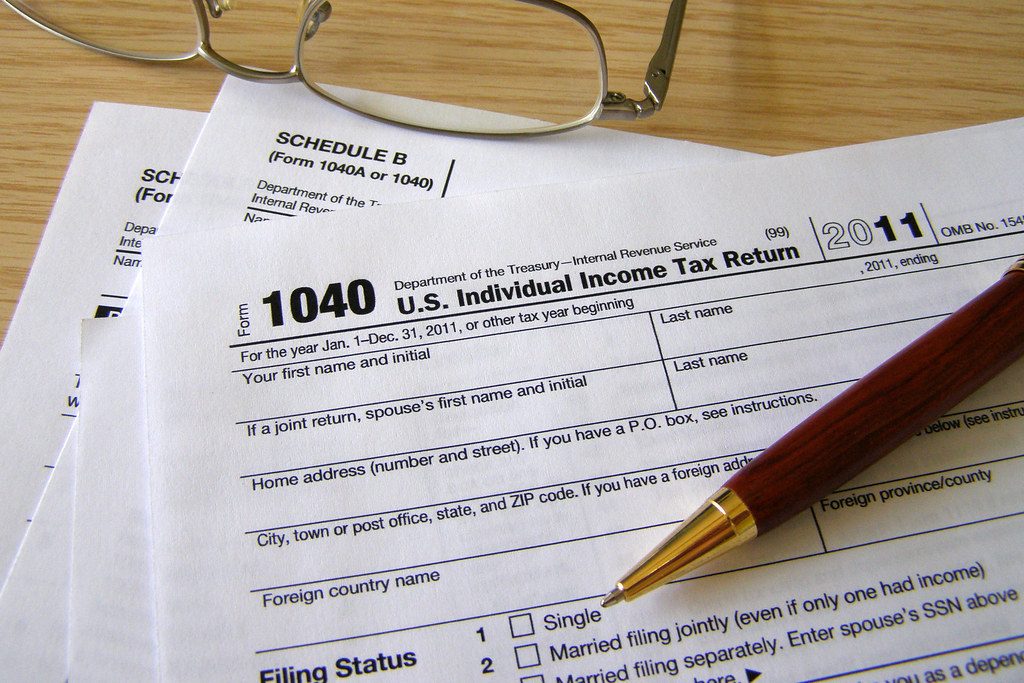 Blank US Form 1040 income tax return pages on a wooden desk, with a wooden ballpoint pen and a pair of reading glasses.