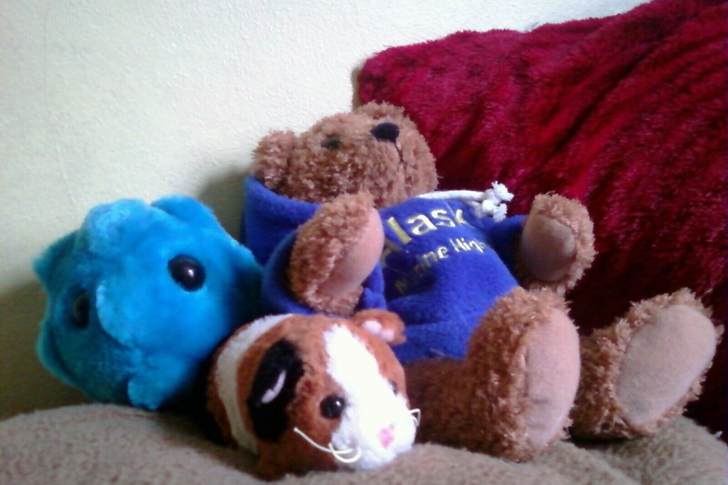 A group of plush toys, including a brown teddy bear in a blue shirt, a blue fuzzy creature and a guinea pig, piled on a sofa.