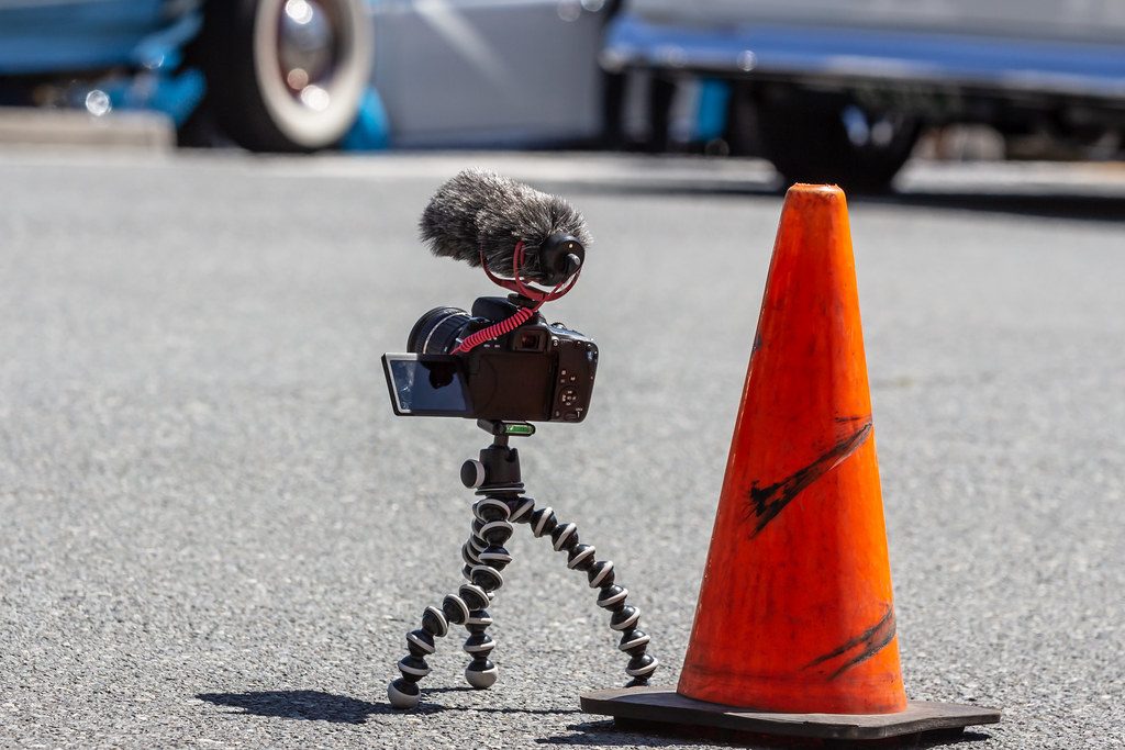 A compact vlogging camera with a flip-out screen and a furry shotgun microphone, mounted on a flexible mini tripod beside an orange traffic cone.