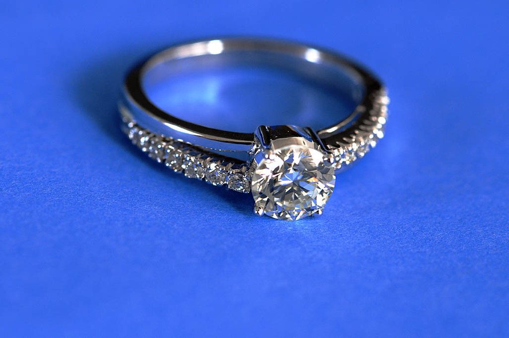 A silver diamond engagement ring with a pavé band resting on a bright blue surface.